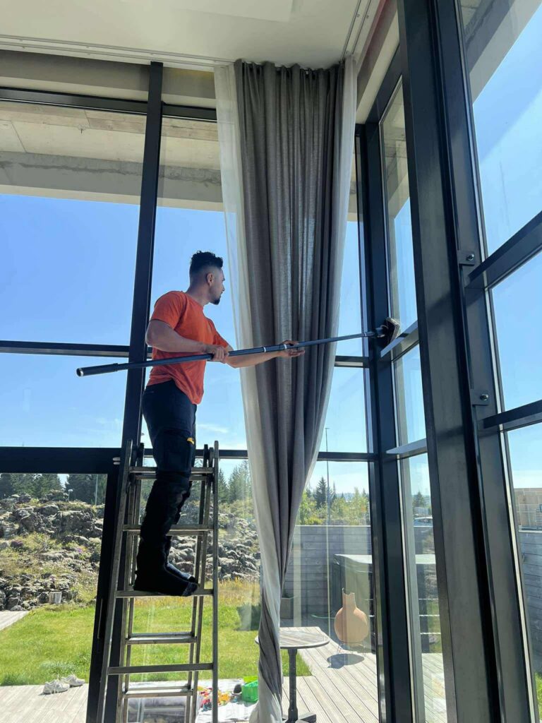 Alt texti: Starfsmaður að þvo háa glugga innandyra með skafti. Fagleg þrif á stórum glerflötum.