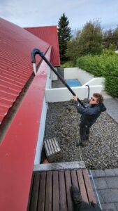 Starfsmaður notar langt ryksuguskaft til að ná upp í rauða þakrennu og hreinsa hana örugglega án þess að nota stiga.