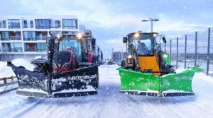Snjómoksturstæki að störfum í íbúðahverfi. Vinstra megin er dráttarvél með svarta Pronar V-skóflu og hægra megin er gul vinnuvél með græna snjóskóflu í snjókomu.