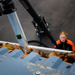 Horft ofan frá í þakrennu fulla af föllnum laufum. Sérhæft ryksuguskaft er komið ofan í til að losa stífluna.