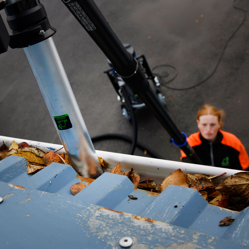 Horft ofan frá í þakrennu fulla af föllnum laufum. Sérhæft ryksuguskaft er komið ofan í til að losa stífluna.