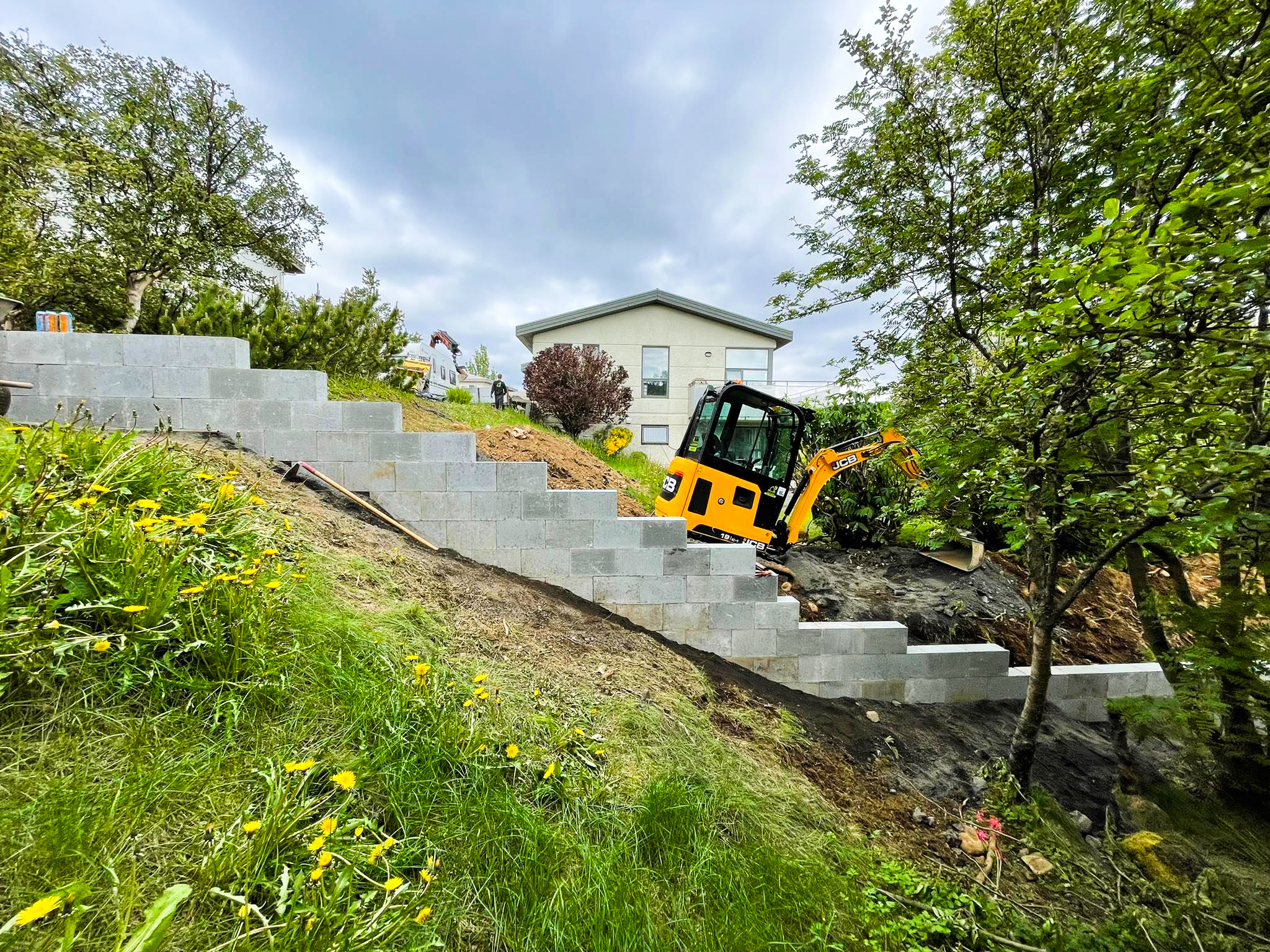 Hlaðinn stuðningsveggur í hallandi garði þar sem unnið er með lítilli gröfu.