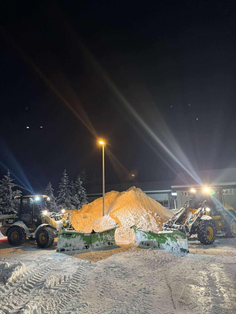 Tvær stórar vinnuvélar með snjótennur að ryðja stórt bílaplan í sameiningu að kvöldi til.