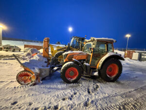 Appelsínugul dráttarvél búin snjóblásara og gul Komatsu hjólaskófla á snævi þöktu athafnasvæði undir flóðljósum.