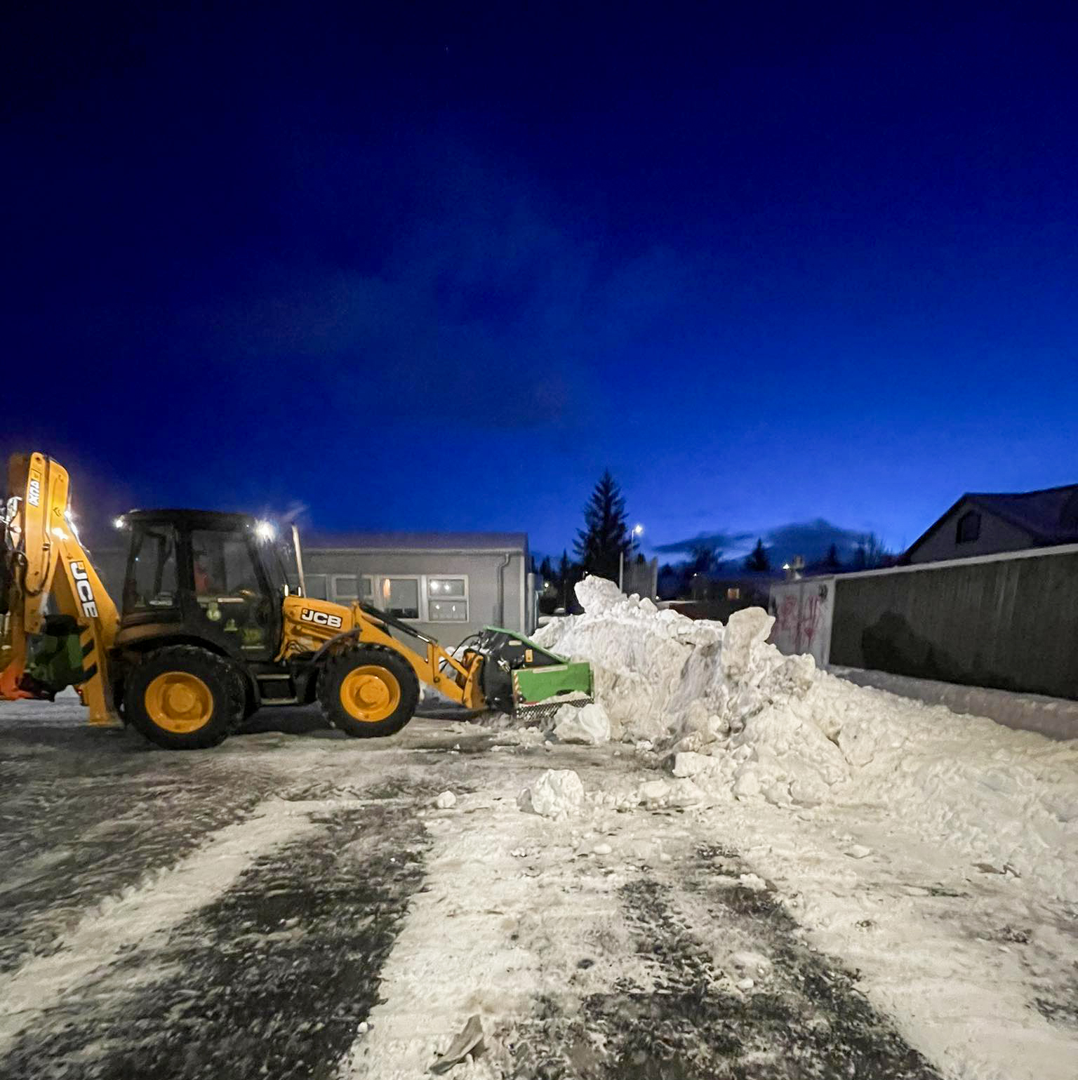 Gul JCB traktorsgrafa að ryðja snjó og búa til snjóhaug í íbúðagötu að kvöldlagi.