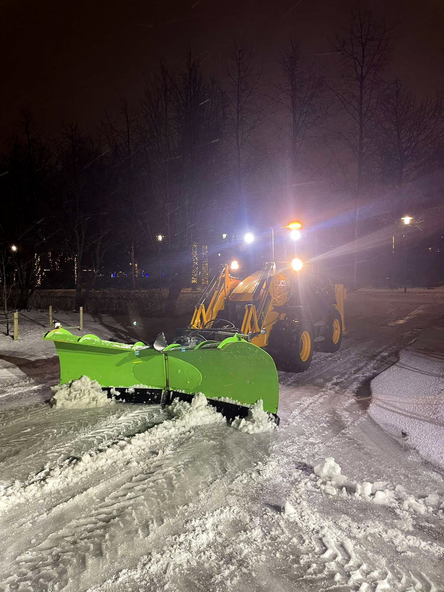 JCB vinnuvél með breiða græna snjóskóflu að hreinsa snjó af göngustíg eða vegi í myrkri.