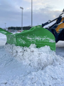 Nærmynd af grænni V-snjóskóflu á vinnuvél að ryðja saman þjöppuðum snjó og ís á stóru plani.