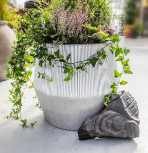 Stór útipottur fylltur með lyngi og grænum plöntum. Við hliðina liggur fallegur útskorinn steinn til skrauts.