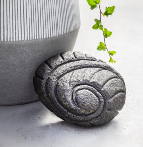 Nærmynd af dökkum steinskúlptúr í formi snigils eða kuðungs, sem skraut við blómapott.