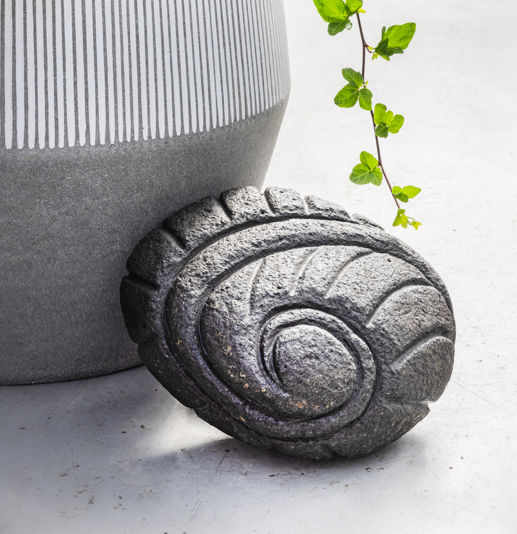Nærmynd af dökkum steinskúlptúr í formi snigils eða kuðungs, sem skraut við blómapott.