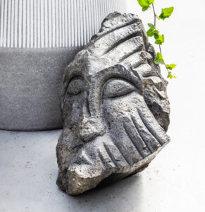 Nærmynd af útskornum gráum steini sem myndar andlit, standandi við hliðina á potti með bergfléttu.