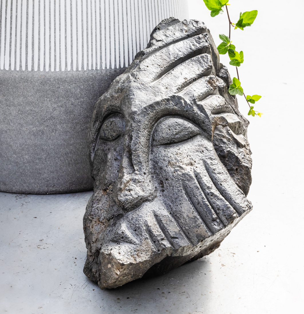 Nærmynd af útskornum gráum steini sem myndar andlit, standandi við hliðina á potti með bergfléttu.