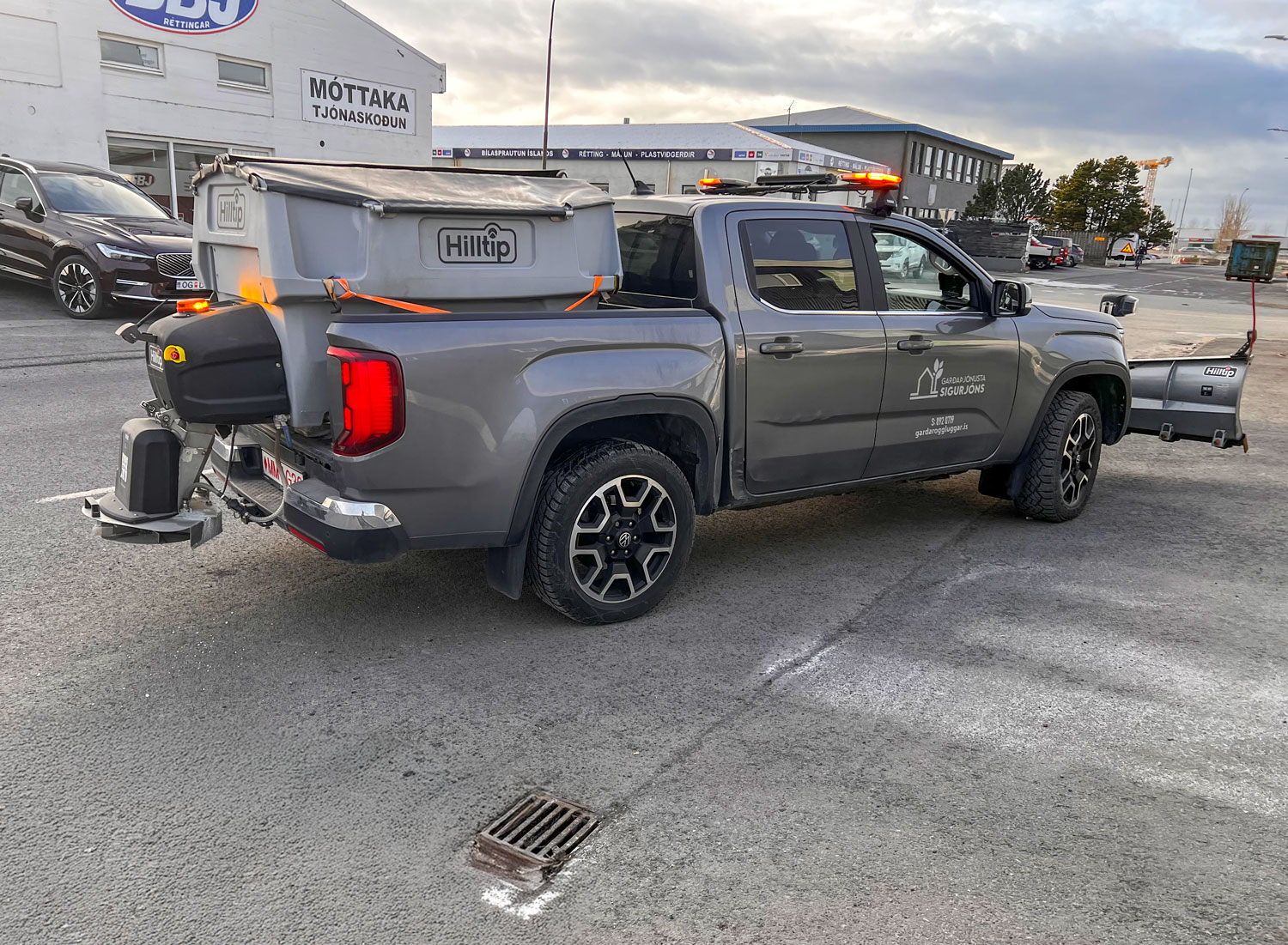 Grár VW Amarok pallbíll búinn Hilltip snjóplóg og saltdreifara fyrir skilvirkan snjómokstur og hálkuvarnir.